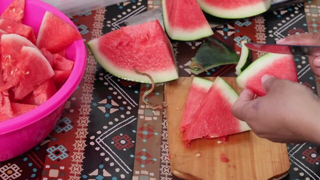 Top view of hands slicing fresh red watermelon into triangles on a wooden board over a patterned tablecloth, preparing a healthy organic snack in the kitchen.
