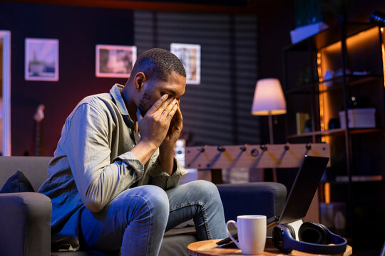 Young black person overworked with a headache in living room, working remotely under stress and pressure from his boss. Young adult frustrated and worried, struggling alone with tasks.