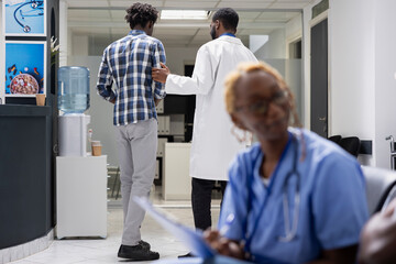 Obraz premium Young male adult follows doctor through hospital hallway for scheduled appointment. Professional physician in lab coat guides patient to clinic office for consultation and medical tests.