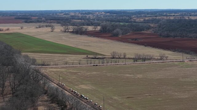 Drone aerial of Oklahoma patchwork farmland with green winter wheat field, red dirt soil, and quiet rural road crossing open agricultural countryside.