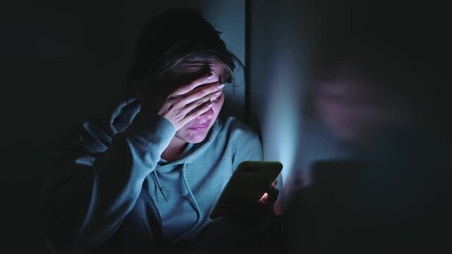 A distressed young woman wearing a hoodie sits in a dark room, reacting to upsetting news on her smartphone and covering her face with her hand, symbolizing digital anxiety.
