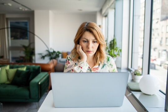 Concerned woman working on laptop at home by window