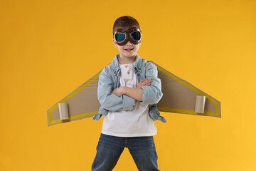 Happy little boy in goggles with cardboard plane wings playing pilot on yellow background © New Africa