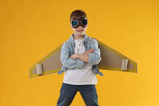 Happy little boy in goggles with cardboard plane wings playing pilot on yellow background