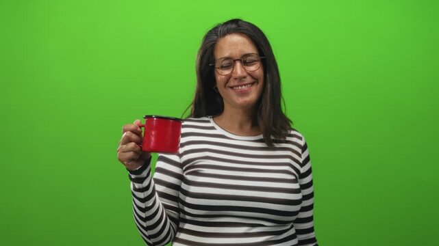 Woman holding red mug up, smiling with one eye closed in green screen studio while wearing striped shirt and glasses; cheerful morning.