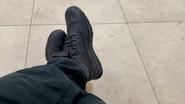 Dirty black boots overhead on tile, worn mens work boots scuffed and dusty, laces tied, dark pants cuff visible, neutral ceramic grout, subtle stain nearby.