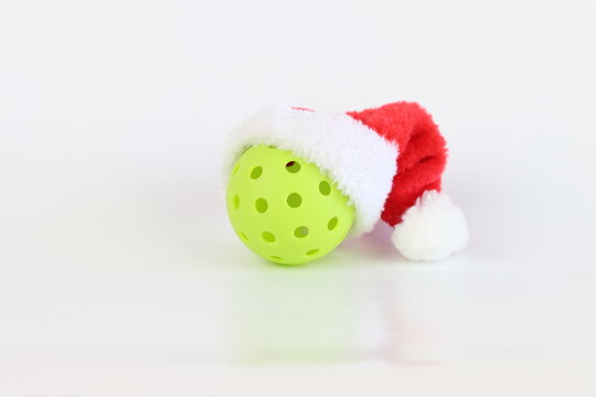 A single lime green pickleball is adorned with a classic red and white Santa hat, ready for Christmas-themed athletic activities