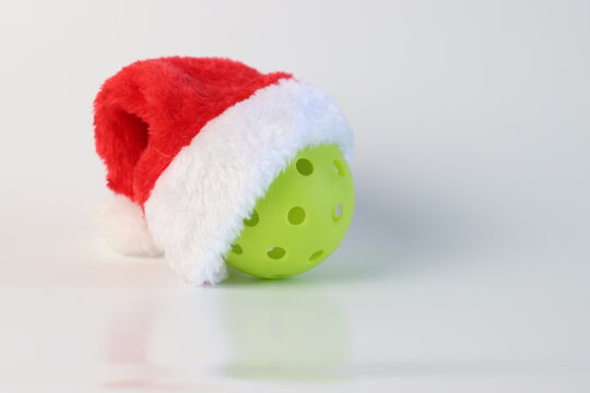 A single lime green pickleball is adorned with a classic red and white Santa hat, ready for Christmas-themed athletic activities