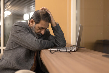 Fototapeta premium Indian tensed senior businessman thinking, worried serious about something while sitting front open portable laptop for client work. long hours of work concept of thoughtful business man