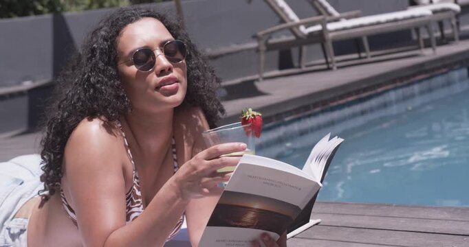 Woman is propping on arms while reading paperback by pool, reaching for drink to cool off