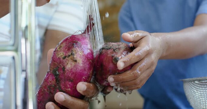 Faucet is running as two hands are rinsing purple yams at kitchen sink for cooking