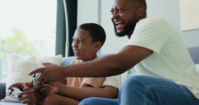 African American dad and teenage son sitting on sofa with white gamepads, reacting to game, bonding