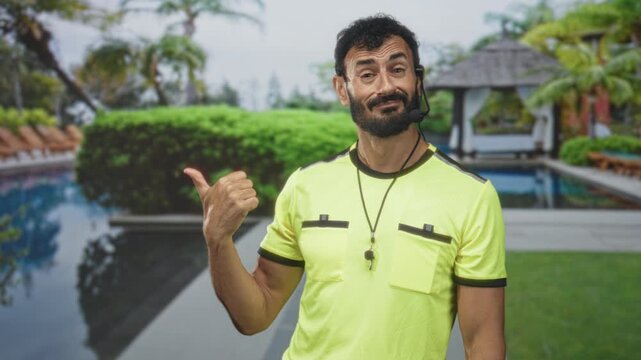 Man referee wearing whistle and headset, pointing thumb at a resort poolside gazebo, visible hand gesture and whistle on lanyard; playful guidance.