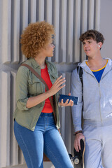 Obraz premium Diverse teenage friends leaning on grooved concrete wall on sidewalk, talking, holding skateboard