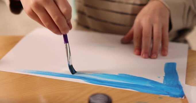 Little boy drawing sky with brush and paints at wooden table, closeup. Camera moving left