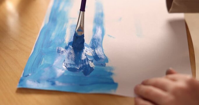 Little boy drawing sky with brush and paints at wooden table, closeup. Camera moving forward