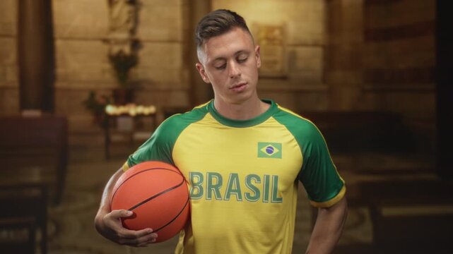 Man holds basketball against hip wearing brasil jersey with brazilian flag patch and eyes closed in building; calm reflection.