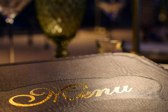 Closeup leather bound menu with golden shiny letters at restaurant table. Fine dining atmosphere with table setting, defocused glassware, ambient lighting, concept of choosing food, hospitality