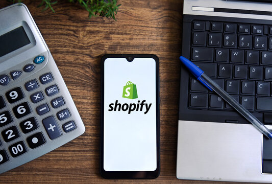 The Shopify logo is displayed on a smartphone screen on a table with a calculator, notebook, and pen. Concept of technology and e-commerce.