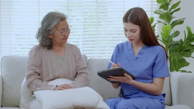 Asian woman caregiver using digital tablet for senior patient consultation, professional nurse checking health record in living room at home, elderly care assessment with checkup, health care support.