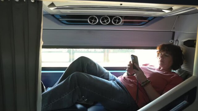 Sleeper Bus Passenger Resting With Device In Cozy Compartment. Teenage Traveler Reclining Comfortably While Browsing Mobile In Softly Lit Sleeping Carriage With Curtains Partly Drawn