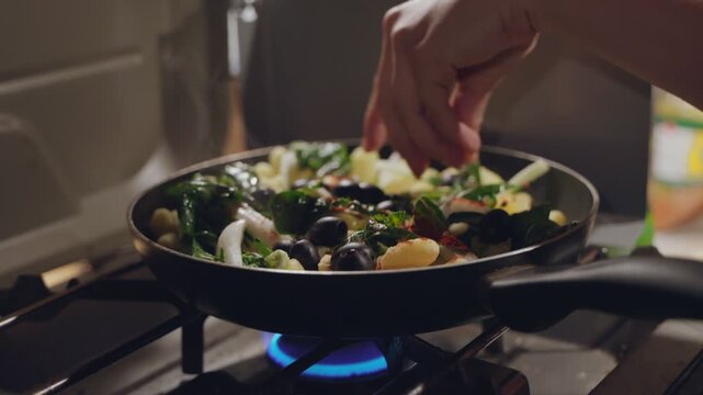 Woman's hand adding olives to gnocchi and greens cooking in a frying pan on a camper van stove in slow motion