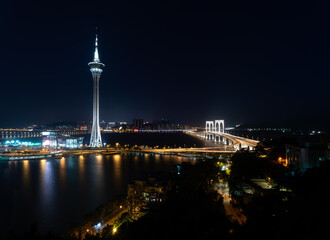 Macau tower and bridge illuminated at night over water.