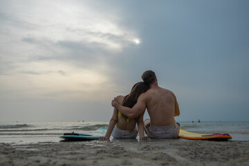 Obraz premium Romantic couple sitting on beach with surfboards at sunset, embracing by the ocean. Summer vacation, love relationship, tropical travel lifestyle and peaceful seaside moment concept.