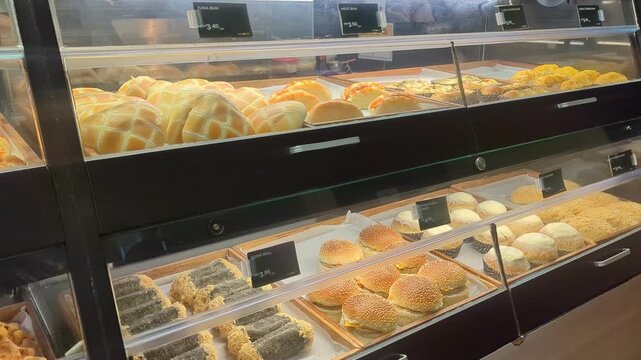Assorted freshly baked buns and pastries displayed on shelves inside a bakery shop glass showcase.