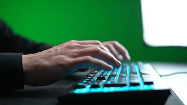 Person typing on a keyboard with blue backlight in a dark room with a green screen