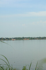 Serene river landscape featuring calm reflective water and distant green tree-lined village under a soft blue sky, evoking peaceful natural beauty.