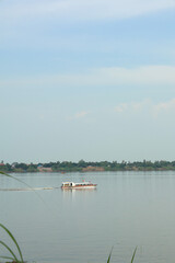 Traditional passenger boat cruising across a calm, wide river under a clear blue sky, showing a distant tropical shoreline.