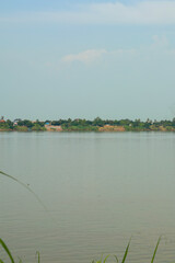 Serene wide river flowing gently with distant village houses nestled among lush green trees under a soft, clear blue sky, peaceful landscape.