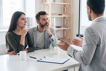 Fototapeta premium Thoughtful young couple listening to financial advisor explaining contract details in modern office