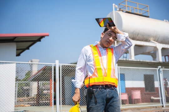 Tired Asian engineer wiping sweat and feeling exhausted while working under the hot sun at an industrial site, Supervisor feeling heatstroke symptoms and resting during a site inspection at plant