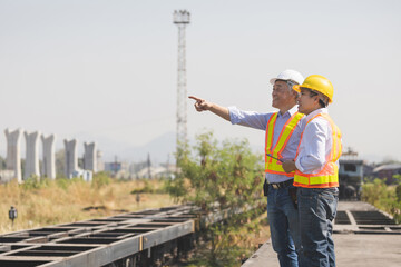 Senior Asian engineers in safety vests pointing and discussing infrastructure development at a railway construction site, Supervisors in hardhats overseeing transport logistics and track construction