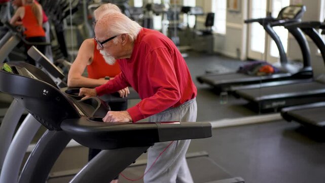 Elderly man engages in workout on treadmill with trainer providing guidance and support in gym setting for healthy activity.