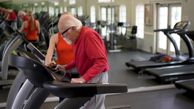 Senior man works out on treadmill with help of trainer in exercise gym, showing dedication to fitness and health.