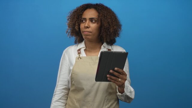 Woman baker wearing apron holding tablet with hand visible in studio against blue backdrop; concentration small business.