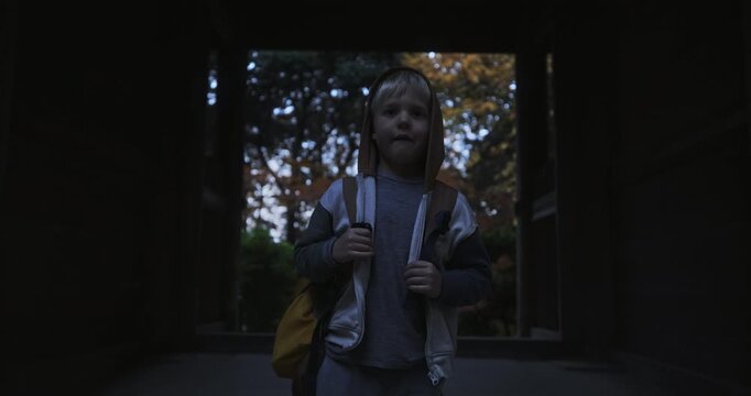 Young boy with backpack standing in shadows of Japanese structure with autumn trees behind - medium shot