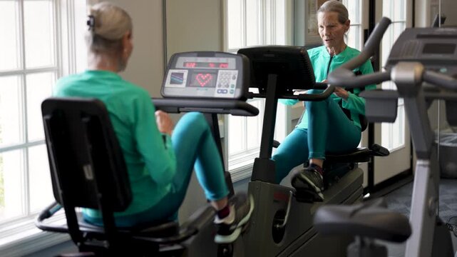 Senior woman works out on a stationary bike in a gym with large windows during her exercise routine for strength and health.