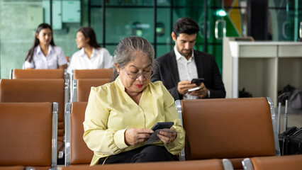 Fototapeta premium Senior woman using smartphone and holding boarding pass while waiting at airport terminal, travel passenger concept, digital check in, modern air transportation, retirement lifestyle
