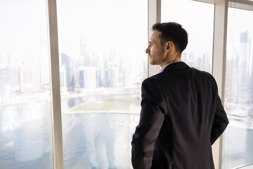 Fototapeta premium Positive thoughtful young property investor man looking at city view out of office window, thinking on real estate project, planning investment, enjoying work break, leisure, daydreaming