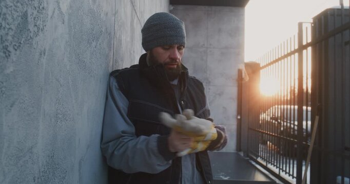 Man Leaning Against Wall Catch Breath After Intense Work. Male Removes Heavy Work Gloves From Frozen, Reddened Hands. Exhaustion, Cold Stress, and Human Recovery Moment After Demanding Winter Labor.