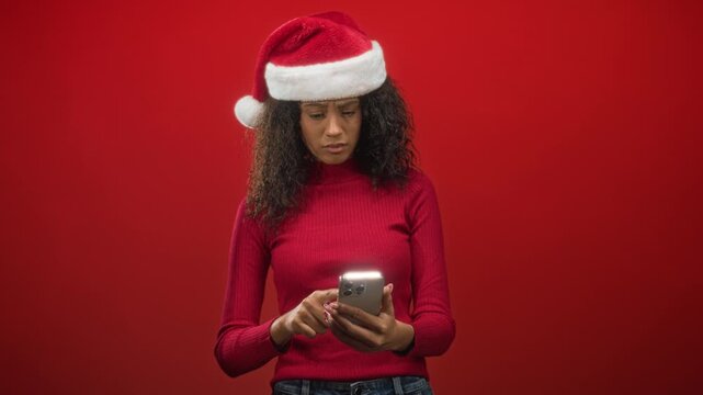 Young woman tapping smartphone with finger while wearing a christmas hat and red sweater in studio; holiday frustration.