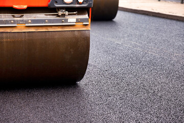 Construction workers are laying asphalt concrete on an urban street. Heavy machinery compacts the fresh asphalt, creating a smooth road surface. The work occurs in broad daylight. © Aleksandr