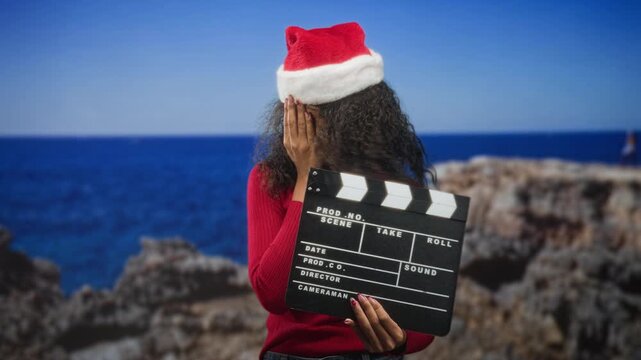 Woman wearing a santa hat holds a clapboard while covering her face with one hand at a studio seaside cliff set; embarrassment holiday fun.