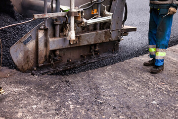 Fototapeta premium Workers are laying fresh asphalt concrete on a city street. Heavy machinery is engaged in the process, creating a smooth surface for vehicles. This urban construction site emphasizes road maintenance.