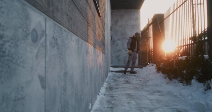 Bearded Man in Beanie And Vest Clears Snowy Path Near Modern Gate. Male Pauses in Work, Inhales Deeply, Leans Against Wall. Domestic Routine, Manual Effort, Winter Weather Resilience in Private Yard.