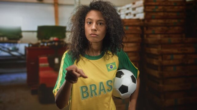 Teenage woman with curly hair wearing yellow brazil jersey holds soccer ball under arm and points finger with exposed hand in a building; determination team spirit.